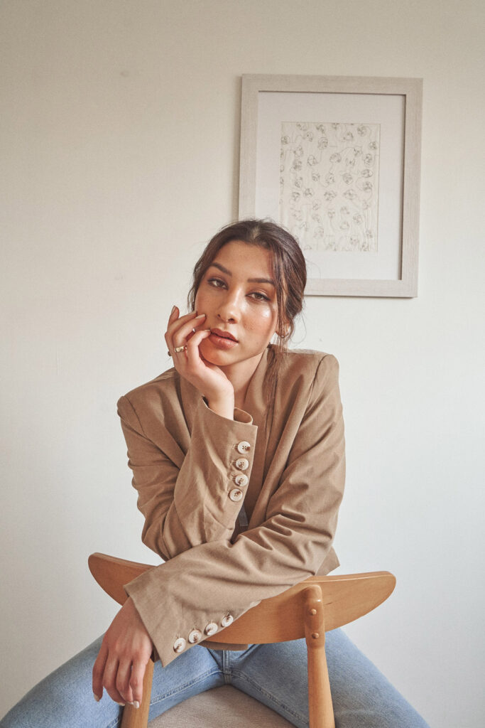 Nadya Inzunza is shown seated on a wooden chair wearing a beige blazer with white buttons and blue jeans. She rests her chin on her hand gazing into the distance with a framed artwork of flowers on the wall behind her.
